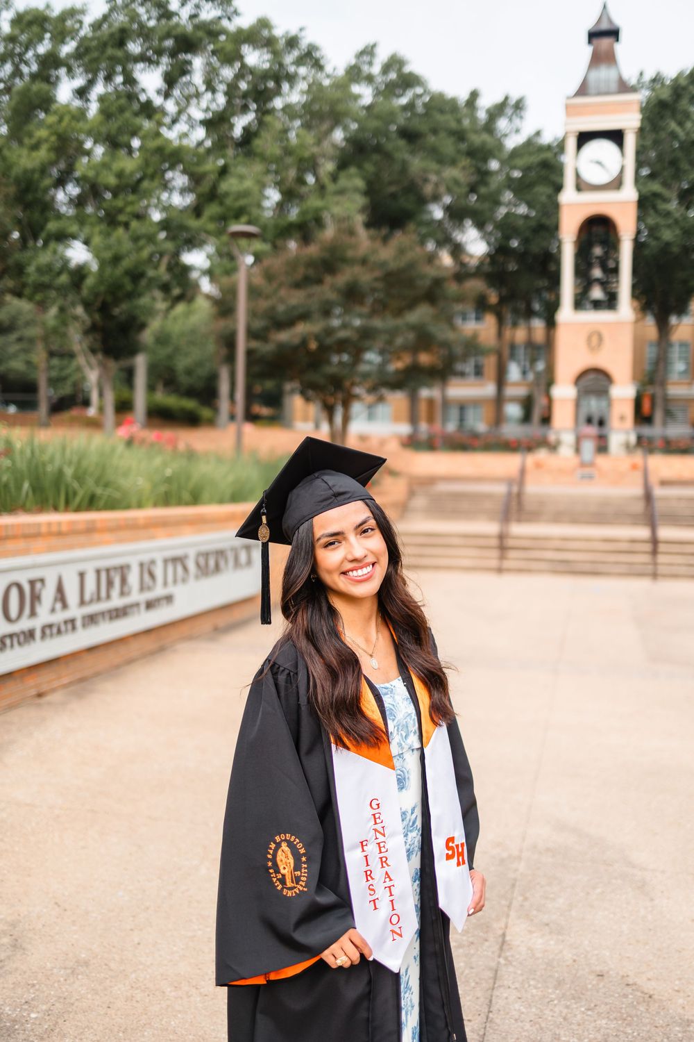 Smiling graduate in black cap and gown with orange stole poses at Sam Houston State University campus.
