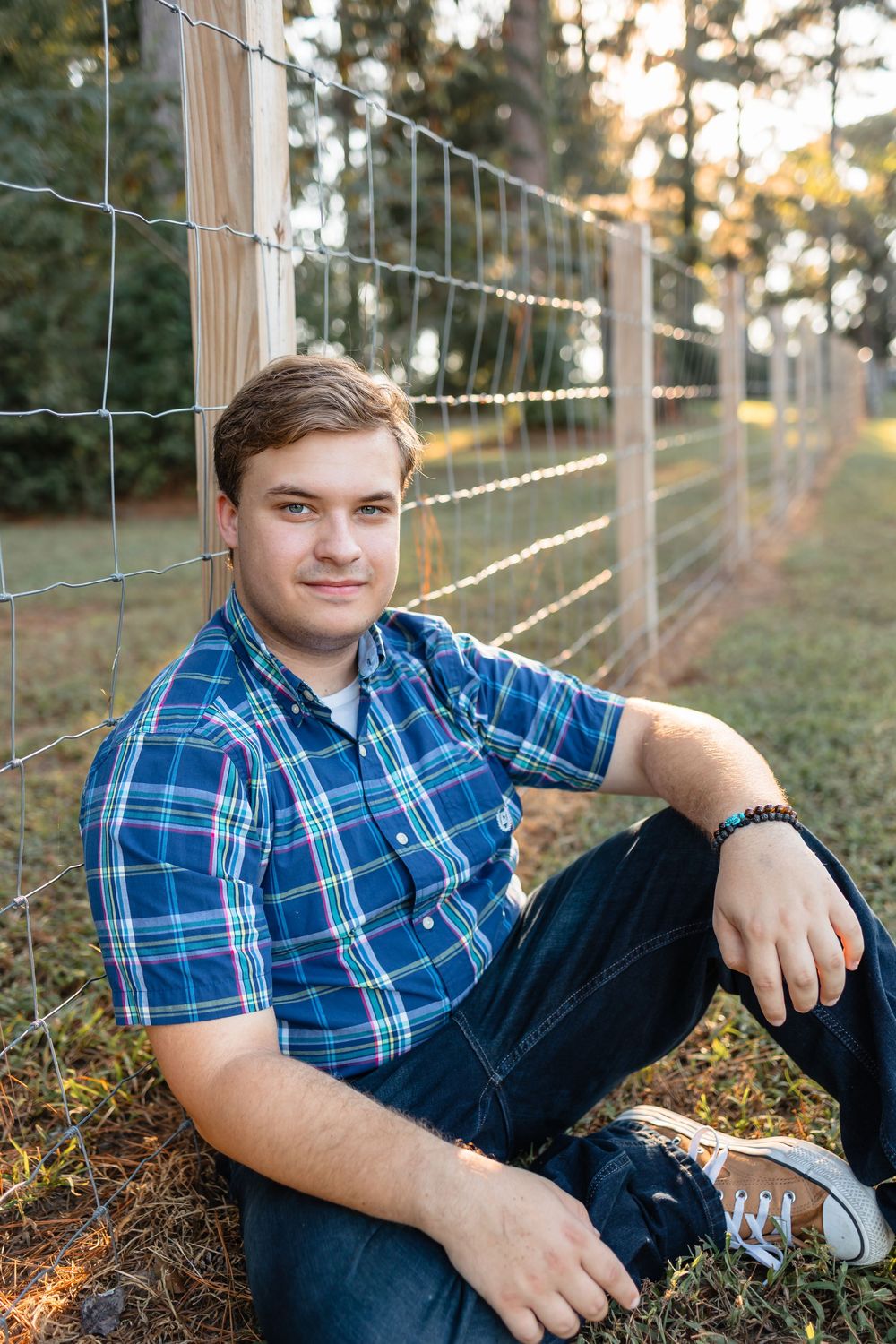 Young man in plaid shirt sitting against a wire fence outdoors at golden hour.