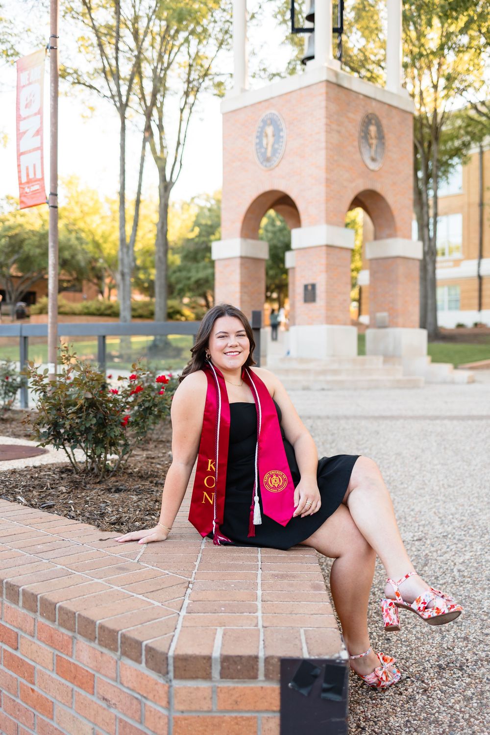 Smiling female graduate in black dress with red sorority stole sits on brick wall near campus arch.