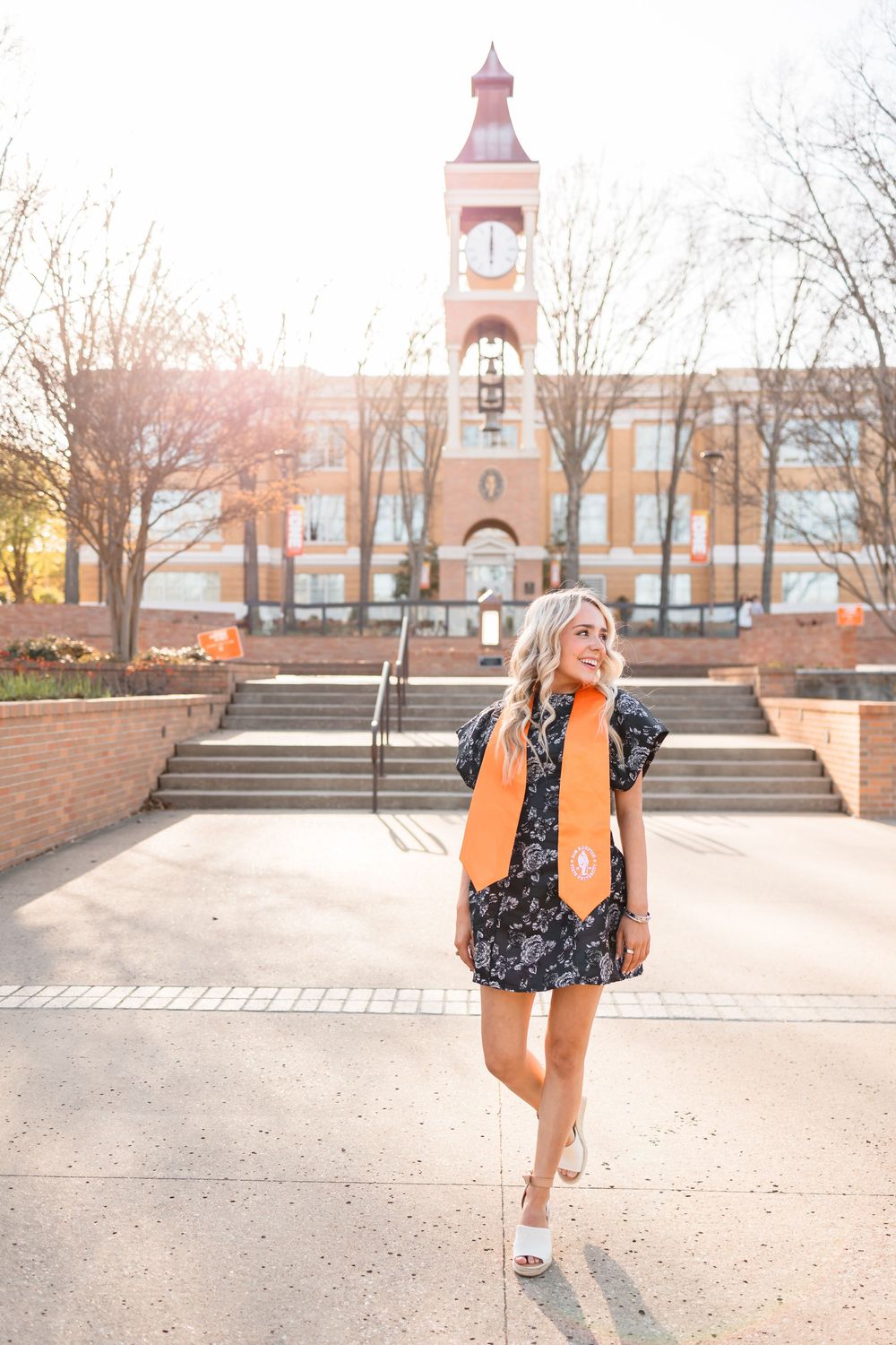 Blonde female graduate in floral dress with orange stole walks near SHSU clock tower at golden hour.