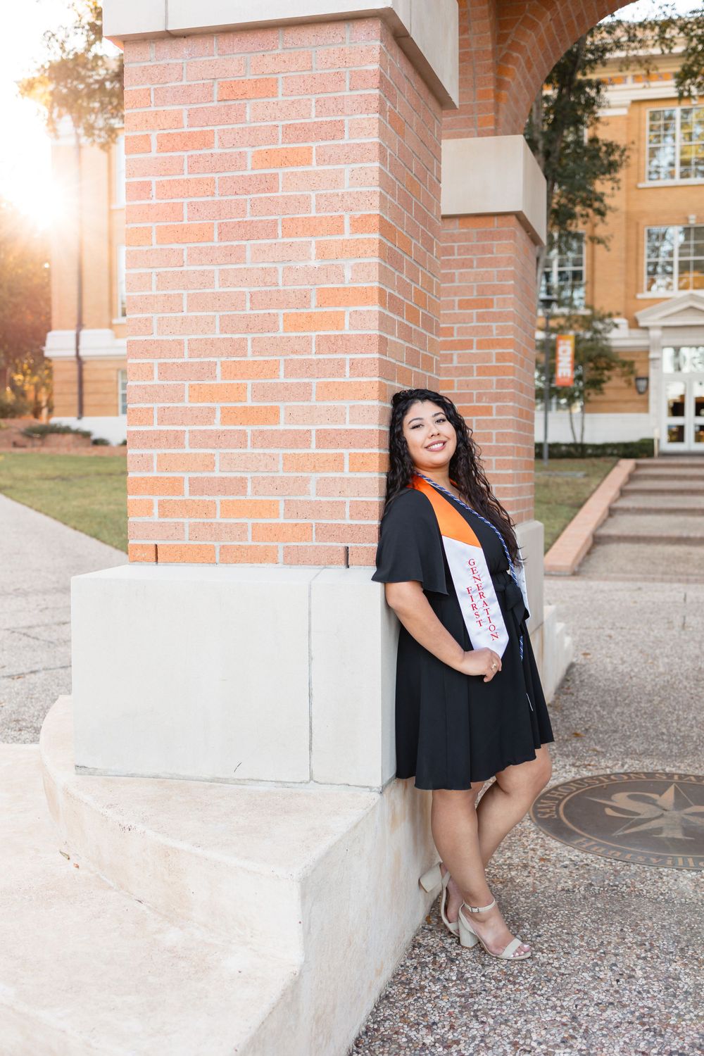 Young Hispanic woman in black graduation gown and white stole leaning against brick column in golden sunlight.
