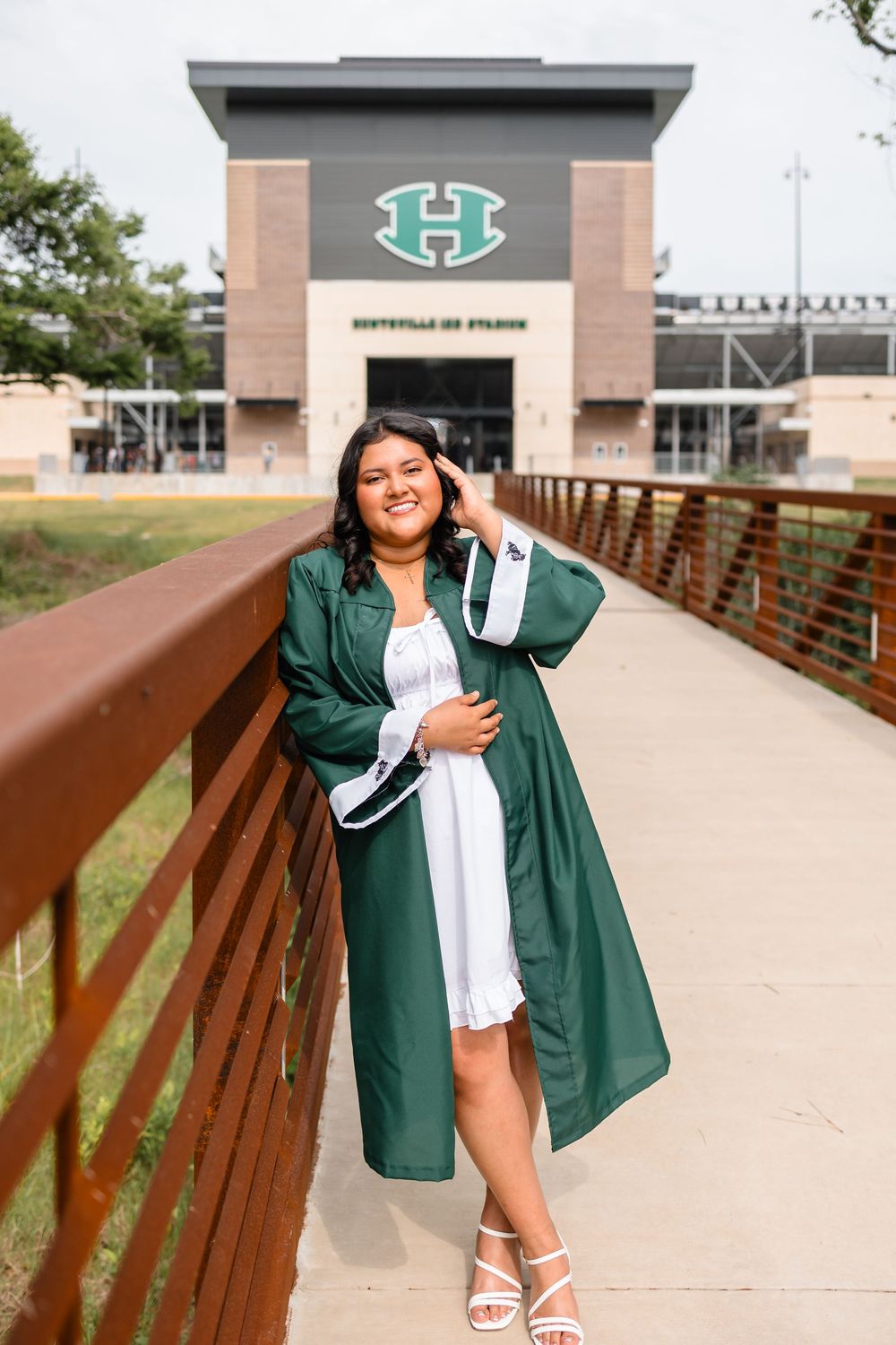 Female graduate in green cap and gown poses on bridge outside Huntsville ISD stadium.