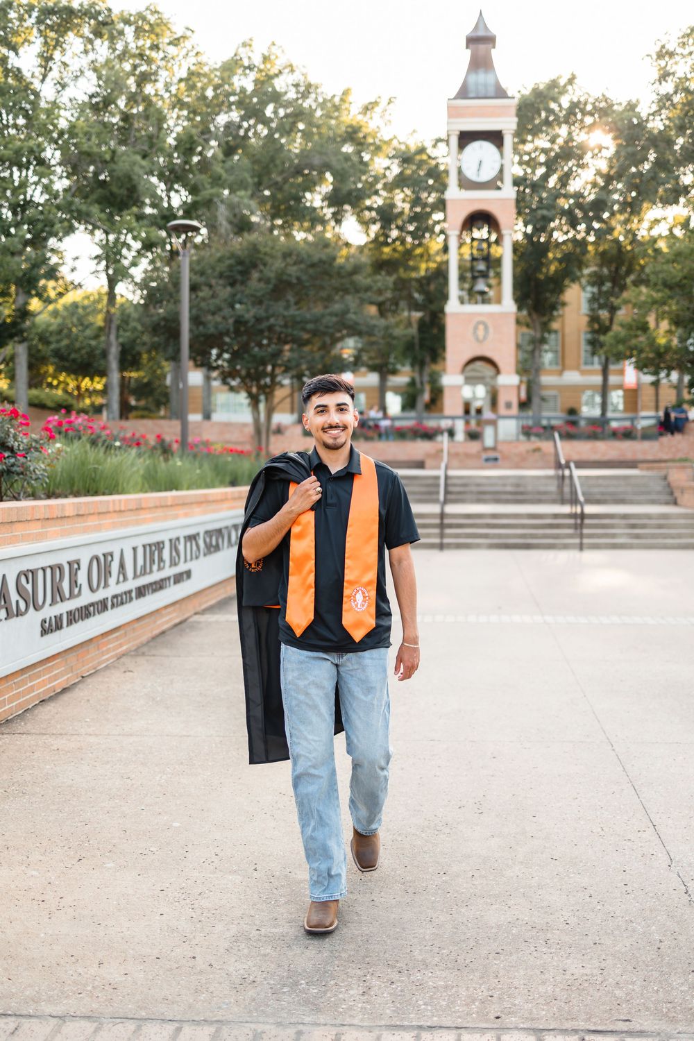 Male graduate carrying black gown walks past Sam Houston State University clock tower at sunset.