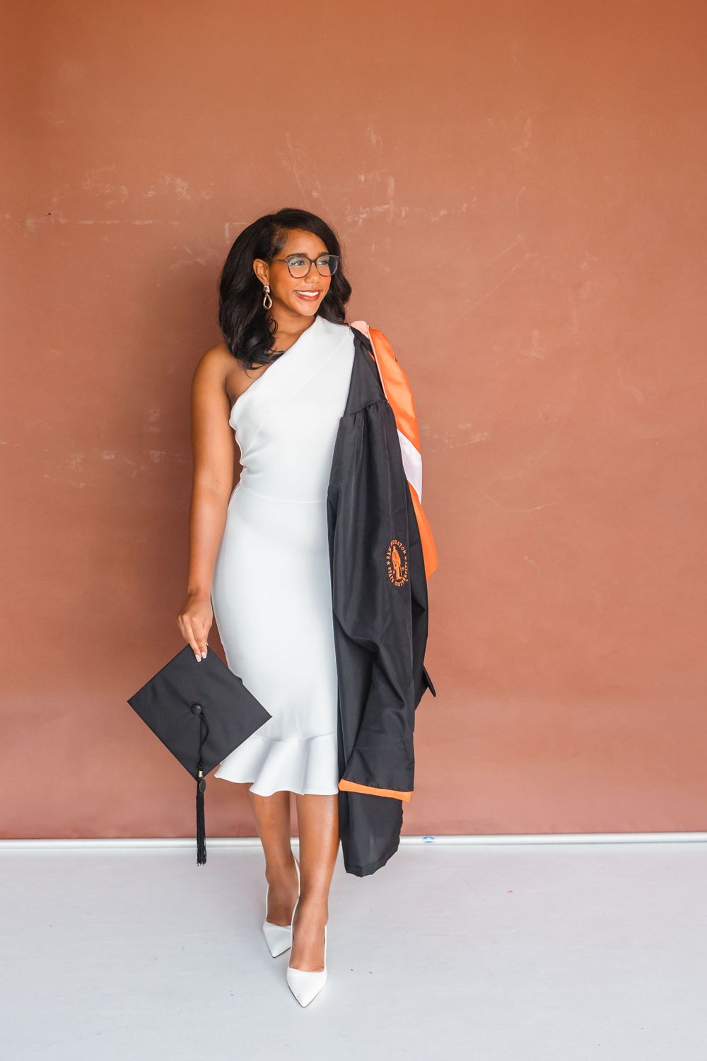 Elegant woman in white one-shoulder dress holding black graduation cap and gown against terracotta wall.