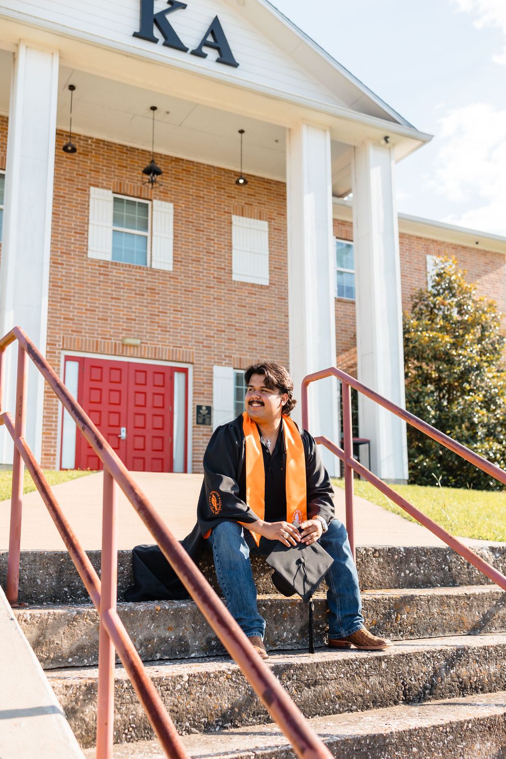 Graduate in orange stole sits on steps of Kappa Alpha fraternity house on college campus.