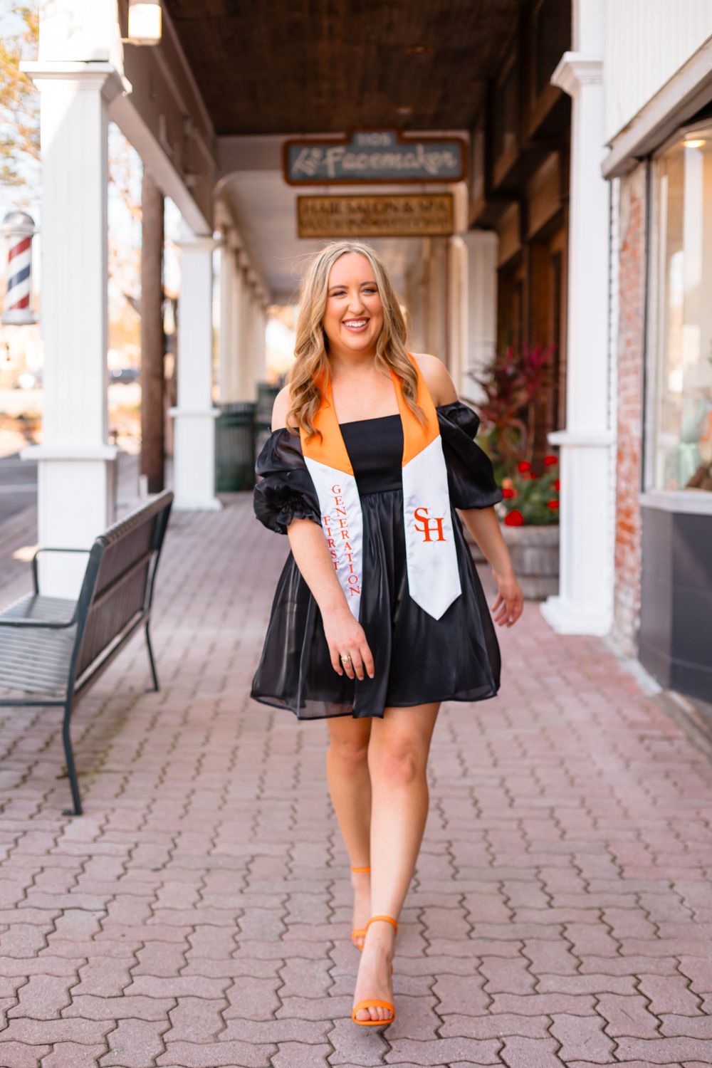 Smiling blonde woman walks in black dress with SH graduation stole and orange heels on brick sidewalk.