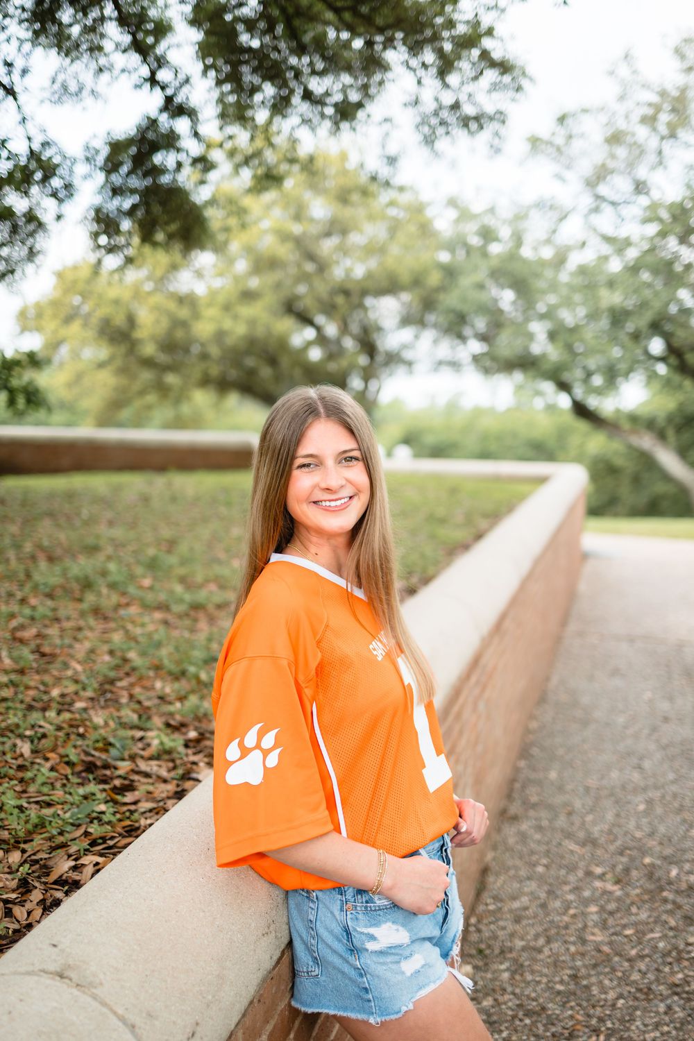 Smiling young woman in orange paw print jersey and denim shorts leans against brick wall outdoors.