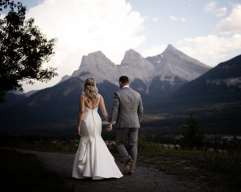Couple in wedding attire walking along trail with mountain vista backdrop.