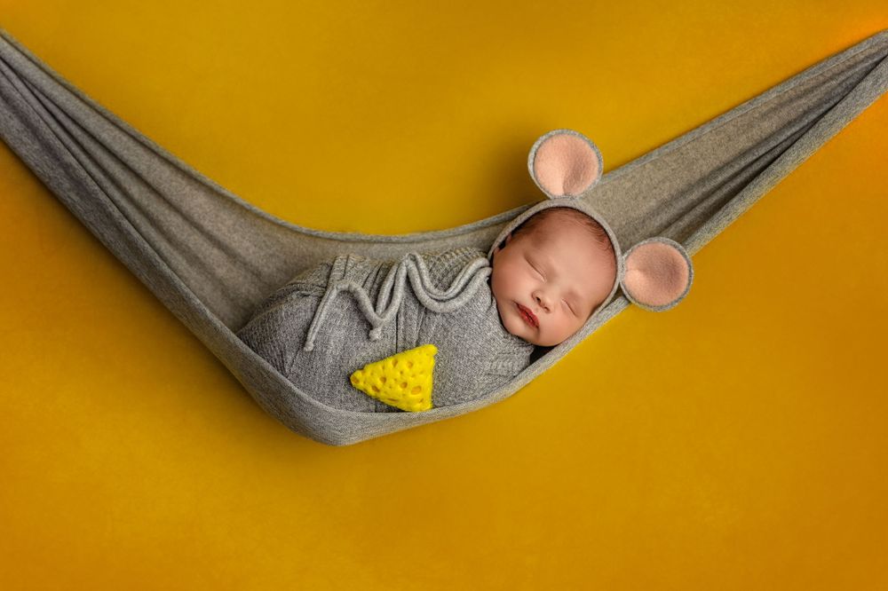 Baby with mouse ears sleeps in gray hammock against vibrant yellow background.