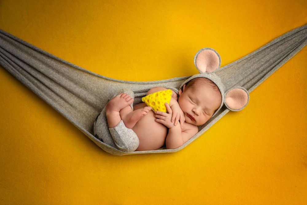 Baby with mouse ears rests peacefully in gray hammock against warm yellow background.