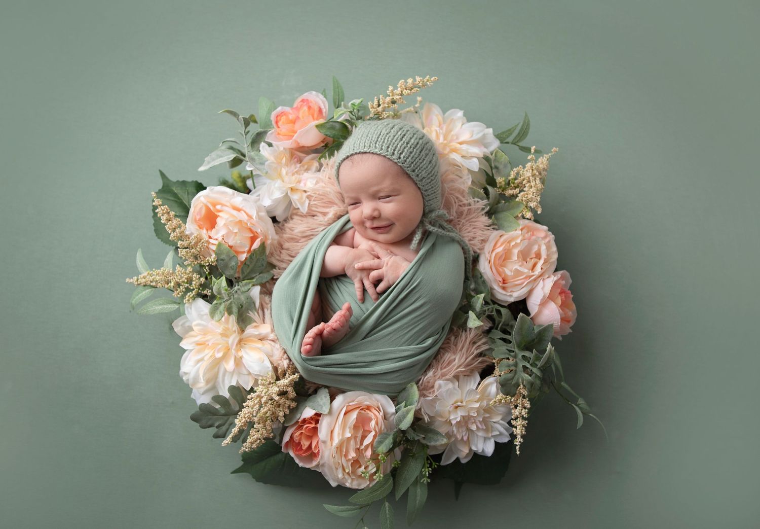 Sleeping newborn in mint wrap surrounded by peach and white flowers on green background.