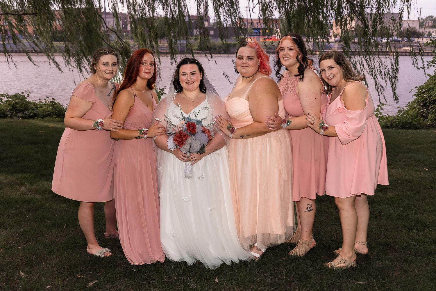 Bridesmaids in coral pink dresses pose with bride in white gown under willow tree.