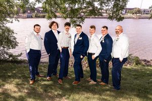 Wedding party in navy suits poses by waterfront under trees.