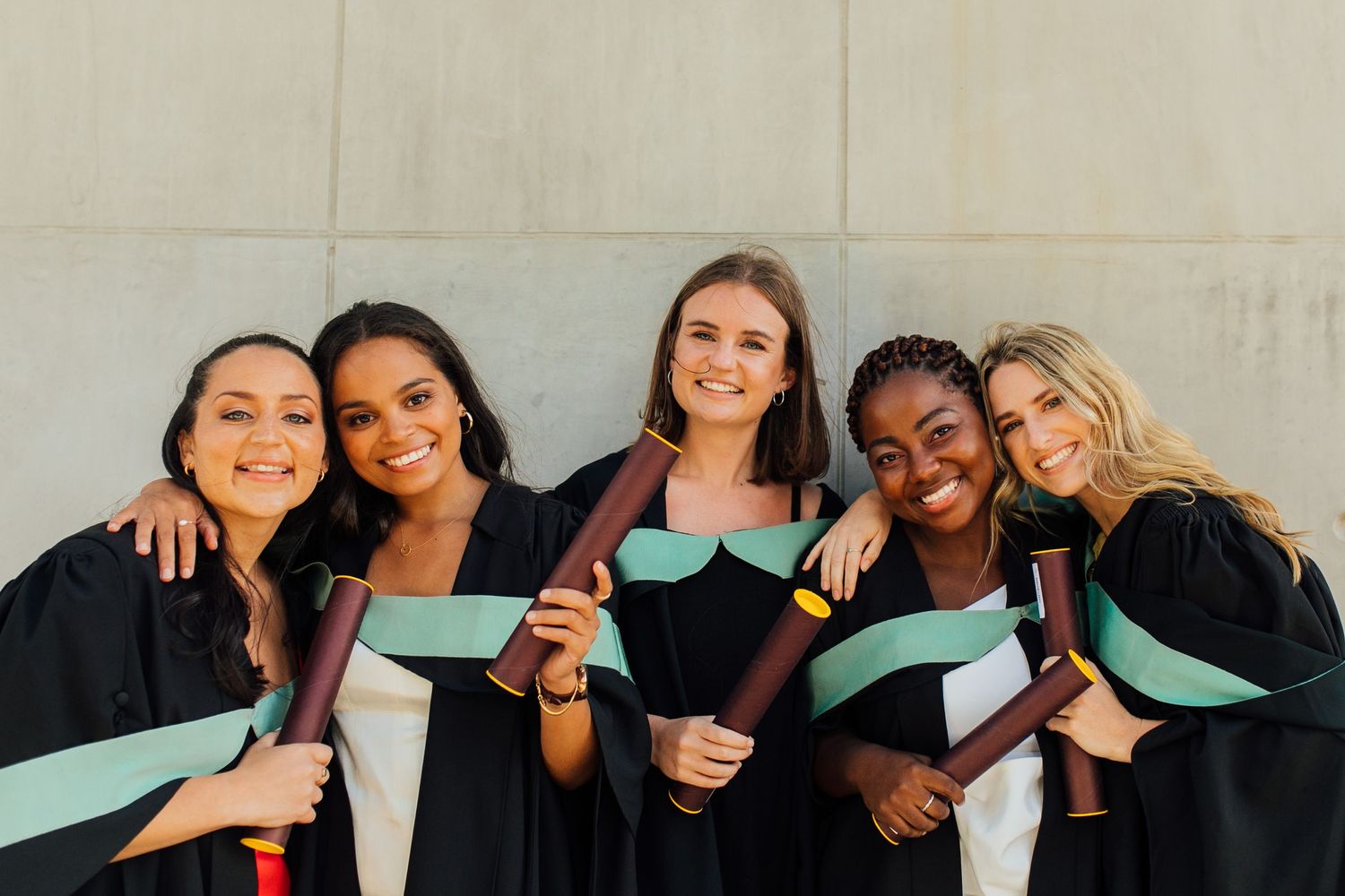 A group of graduates in black academic gowns wearing mint green stoles smile together while showing their diplomas.