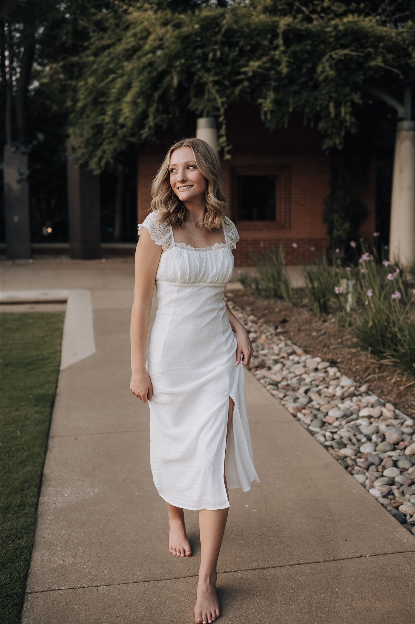 Someone in a white summer dress walks barefoot along a concrete path near a garden.