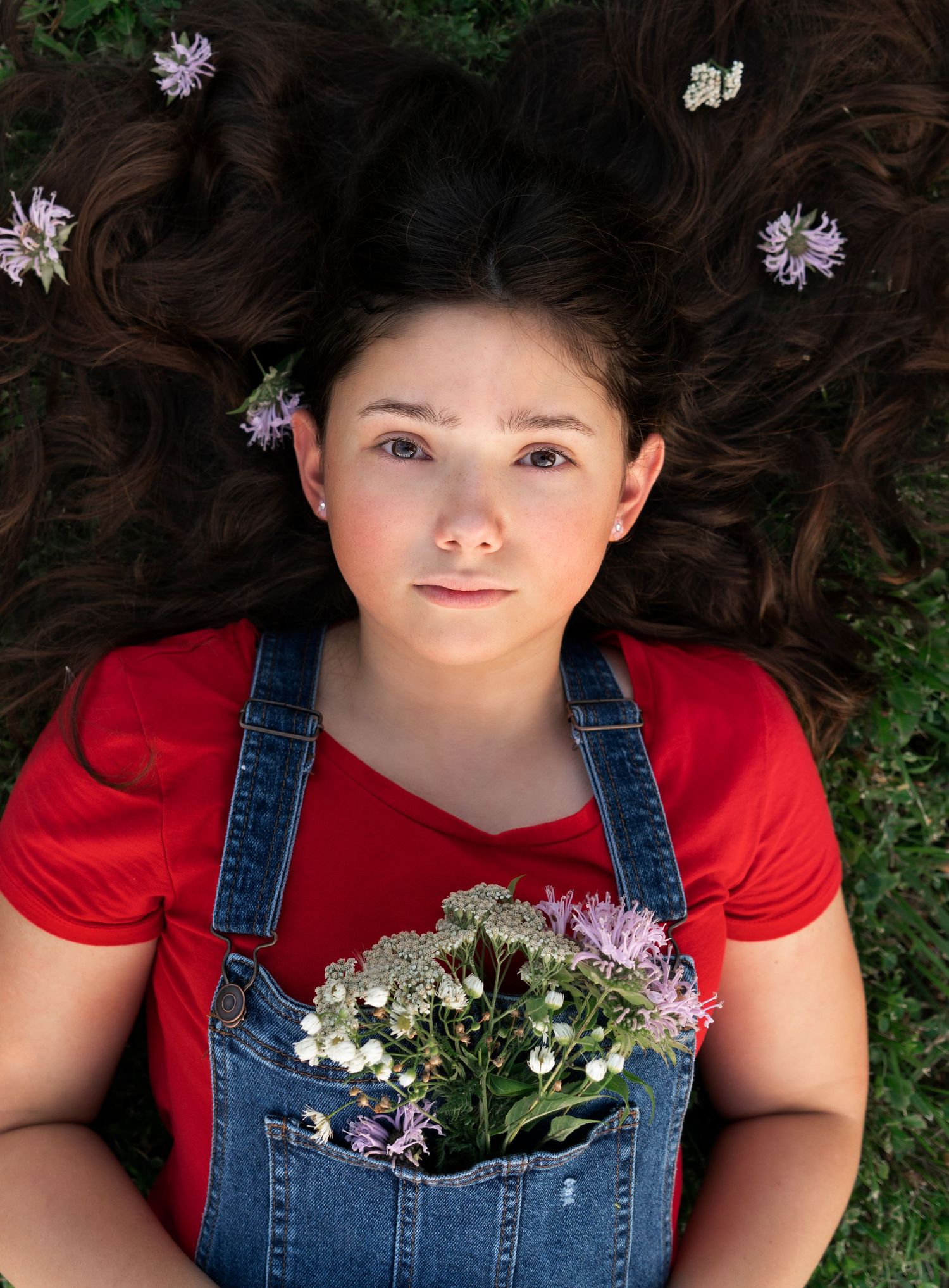 teen girl lays in the grass, looking up at camera with flowers in hair