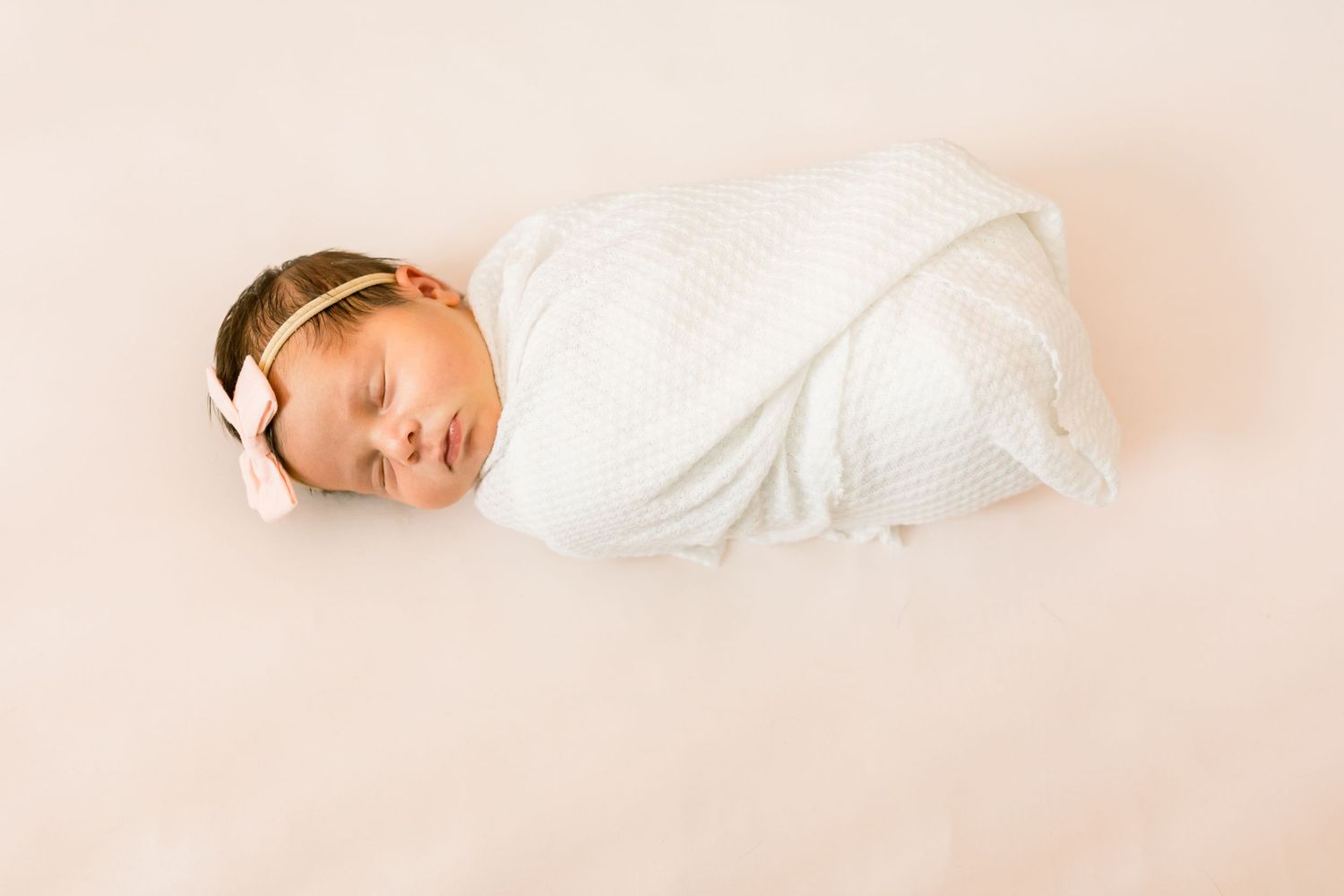 Swaddled newborn with delicate headband resting on soft pink background.