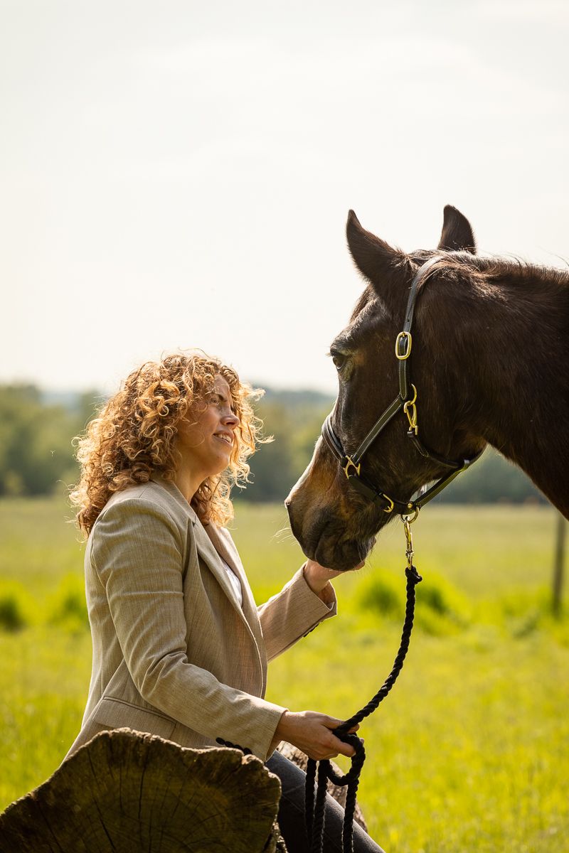 A woman in a beige jacket bonds with her bay horse in a sunlit meadow.