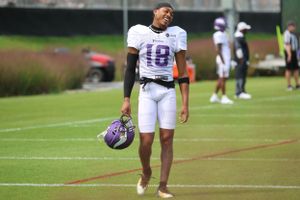Football player in white uniform and purple helmet standing on practice field.
