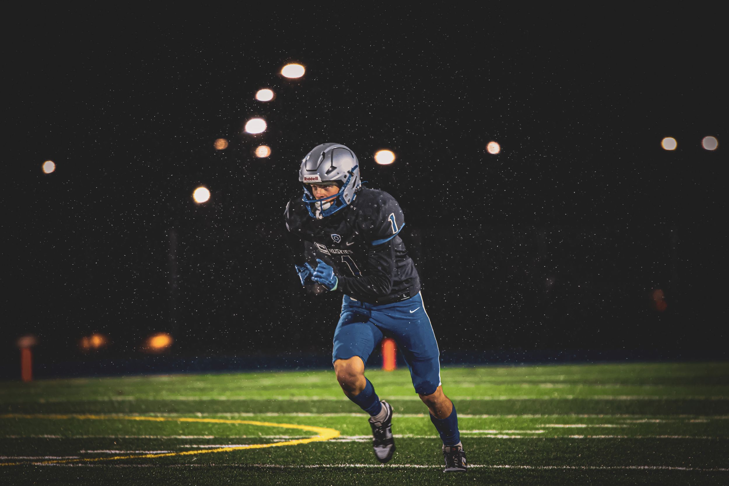 Football player in blue uniform runs across field during nighttime game with stadium lights visible.