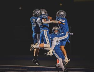 Football players in blue uniforms celebrate with an energetic jump celebration during a nighttime game.