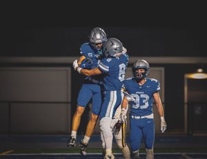 Team members in blue football uniforms celebrate together on the field at night.