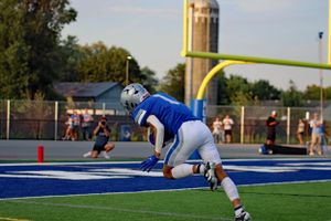 Football player kneeling on blue turf field with goal posts in background.