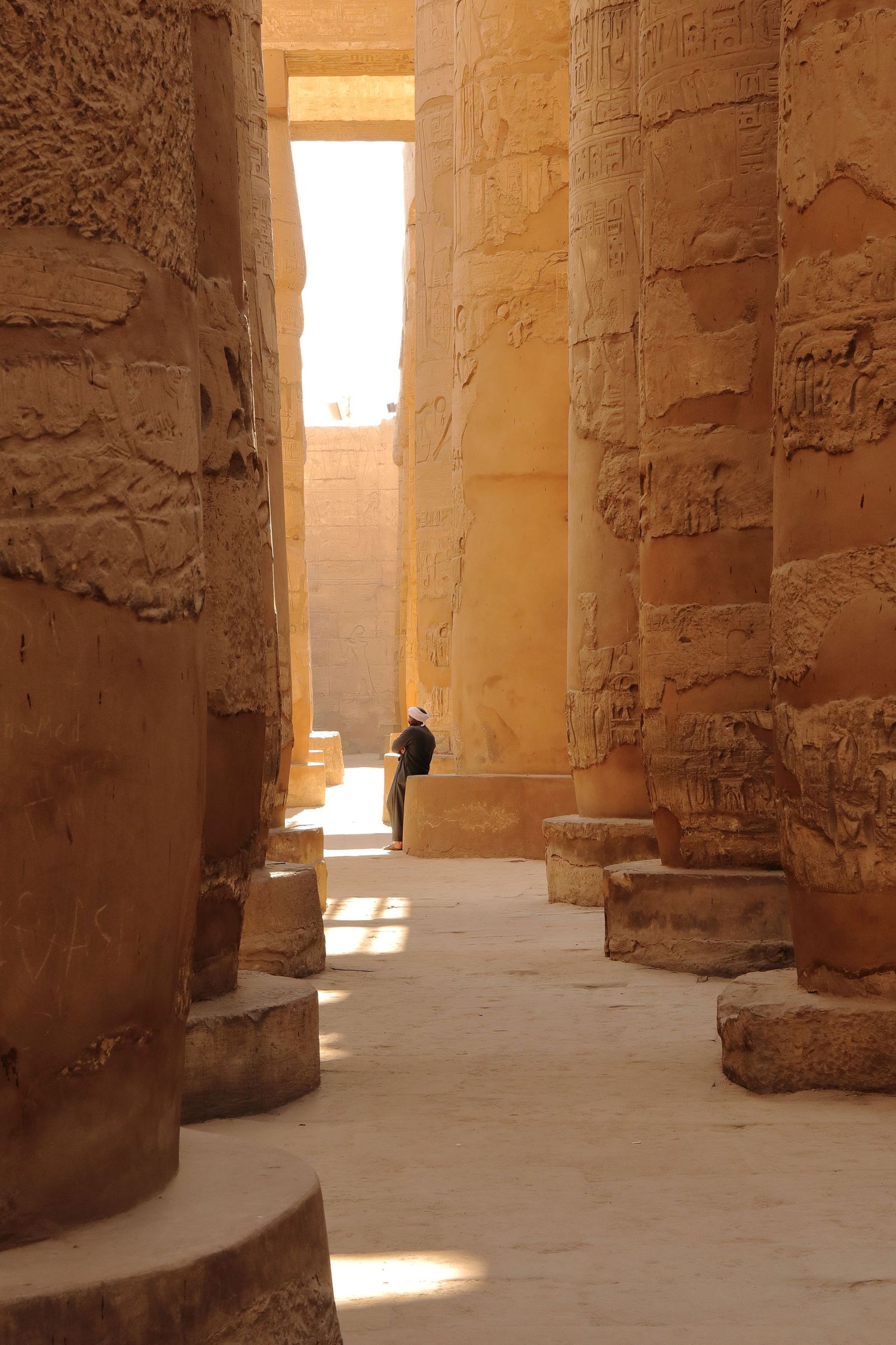 Sunlit corridor between towering columns in ancient Egyptian temple with distant figure.