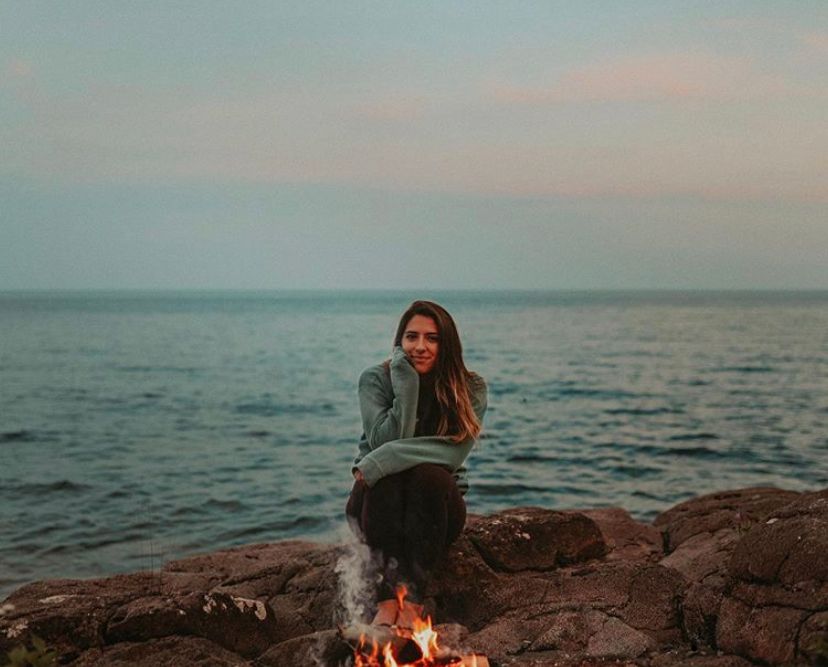 Person sitting by a campfire on rocky beach at sunset with ocean in background.