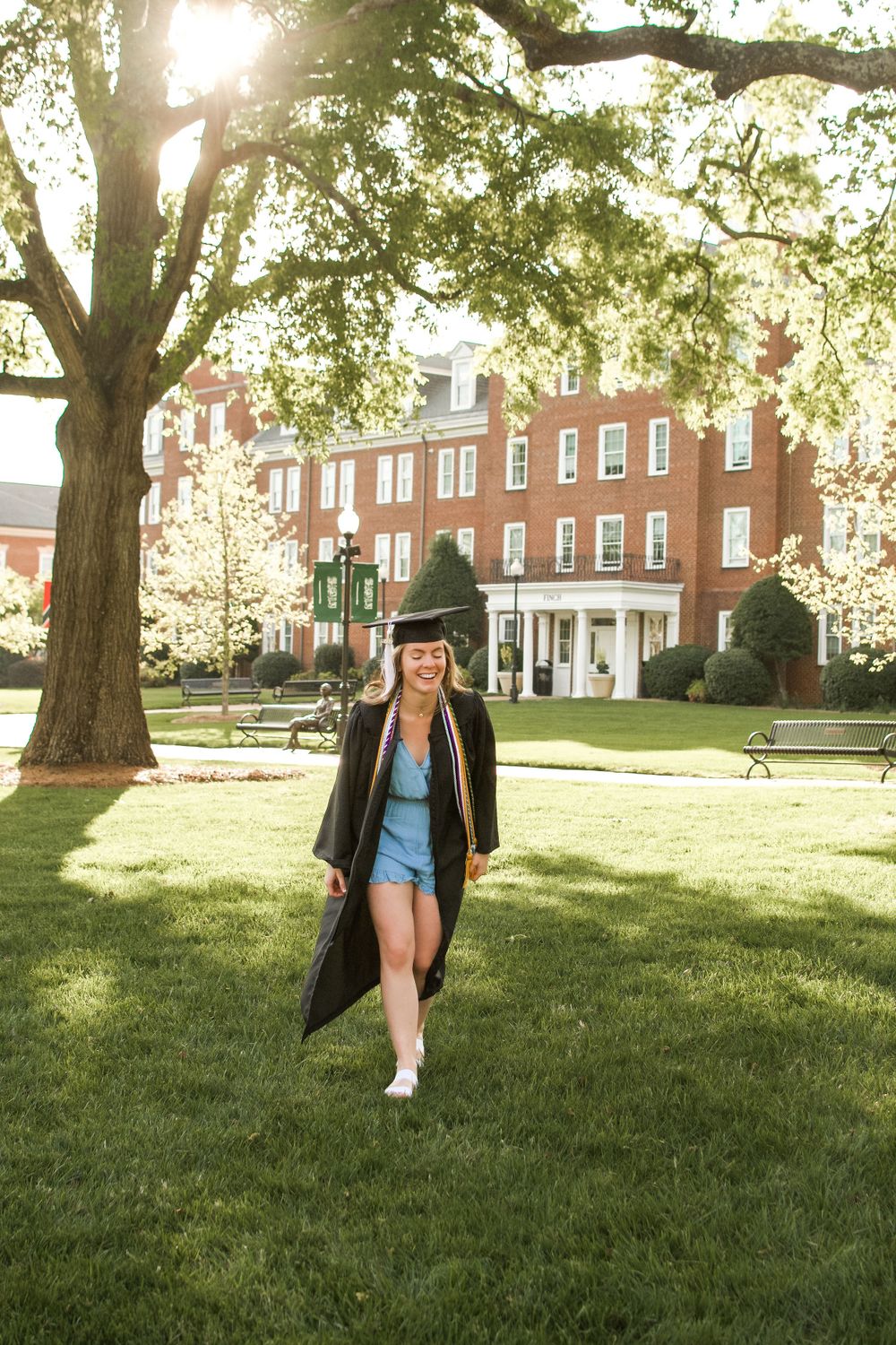 A woman is on a college campus wearing her graduation cap and gown. She is walking and laughing.