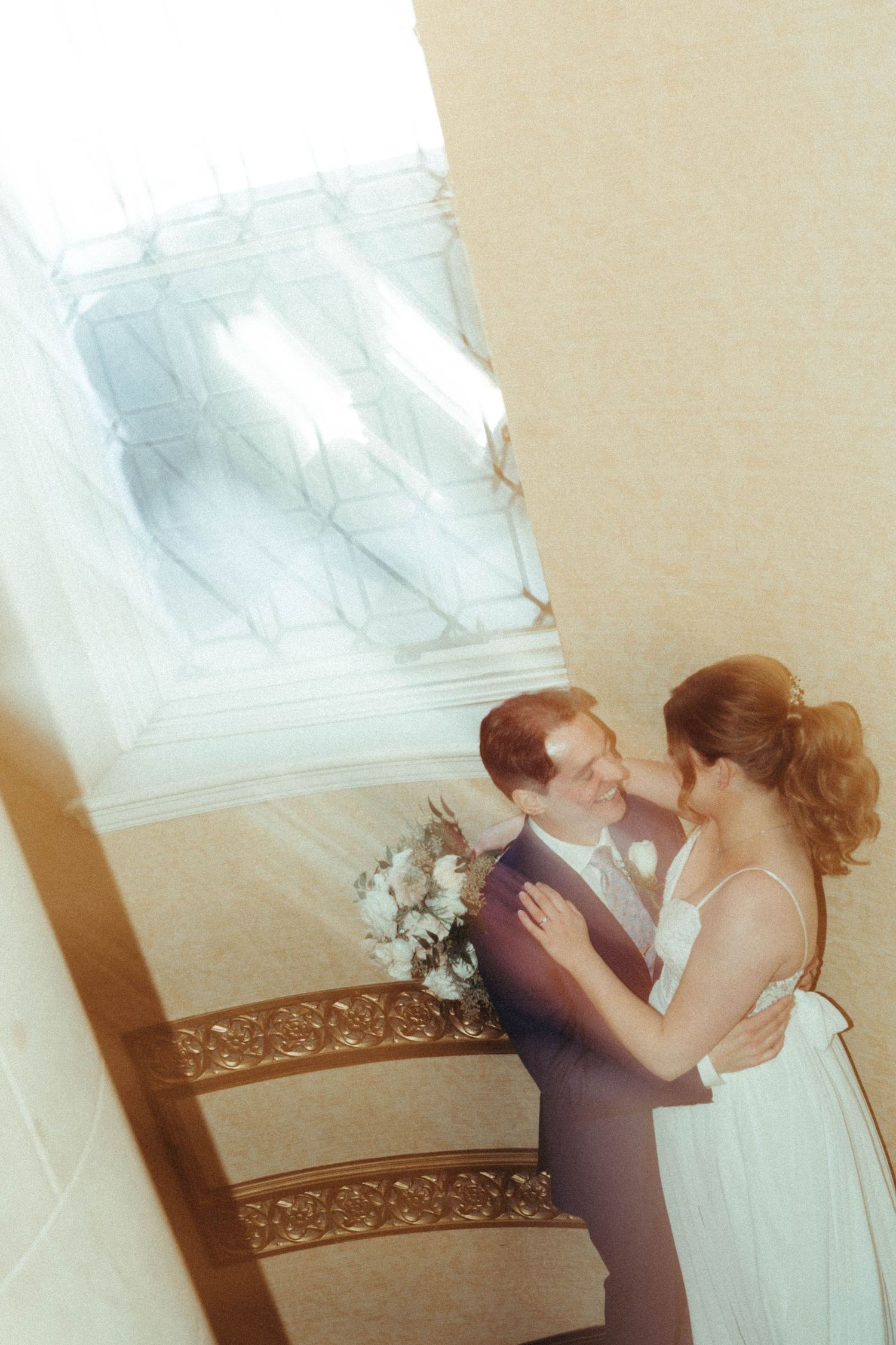 Romantic wedding photo of couple embracing on ornate staircase with sunlight streaming through window.