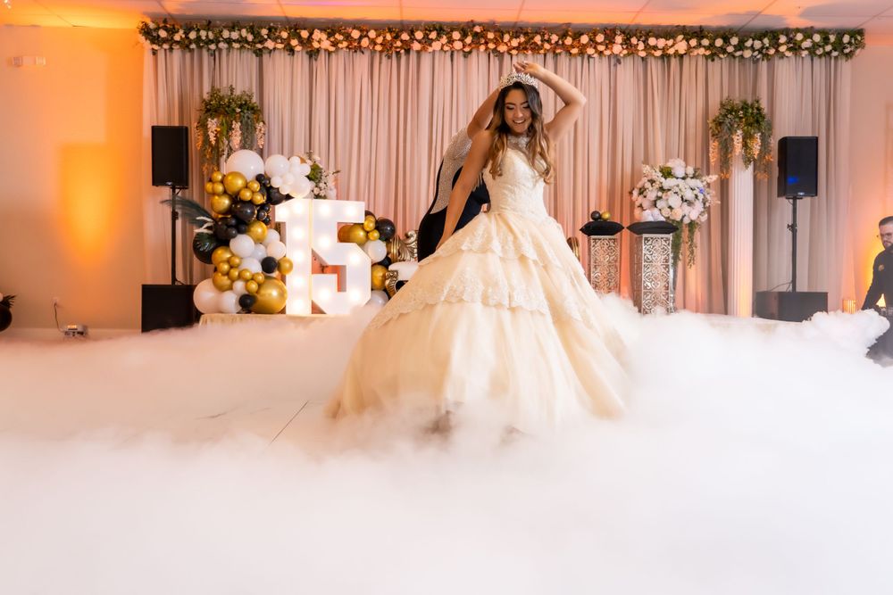 A bride in a voluminous ballgown twirls on a white dance floor with warm lighting and floral decor.