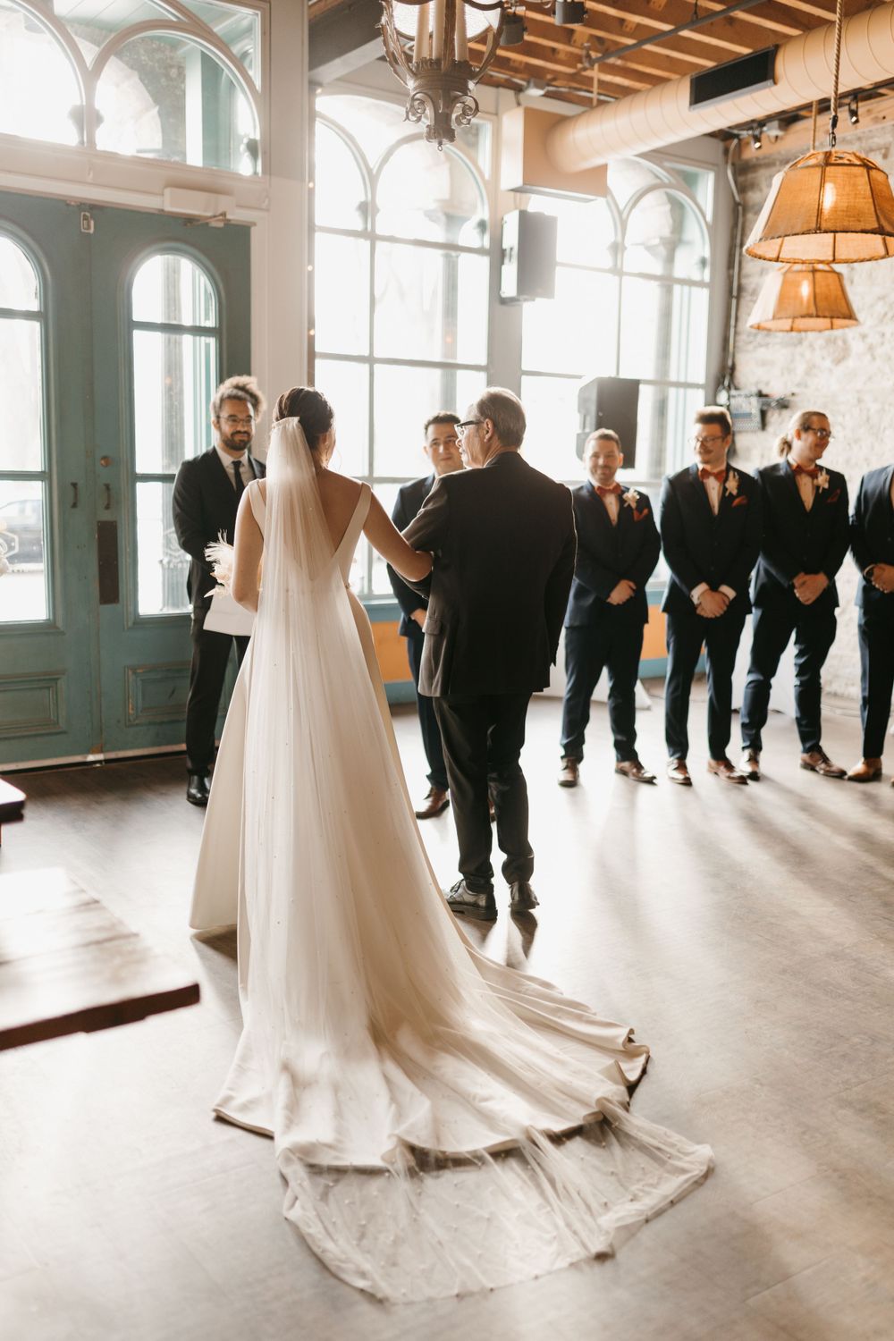 Bride in elegant white gown with long train standing at altar with wedding party in bright venue.