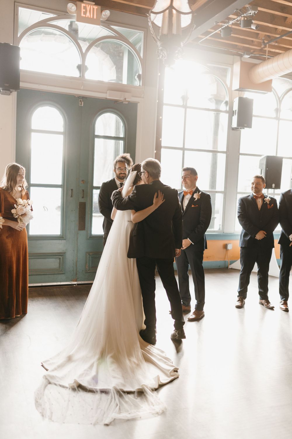 Bride and groom sharing first embrace at altar with wedding party watching in sunlit elegant venue.