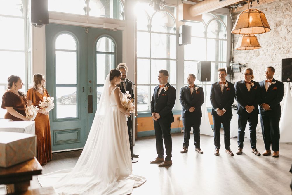 Bride in white gown facing groom and groomsmen at altar near teal arched doors during ceremony.