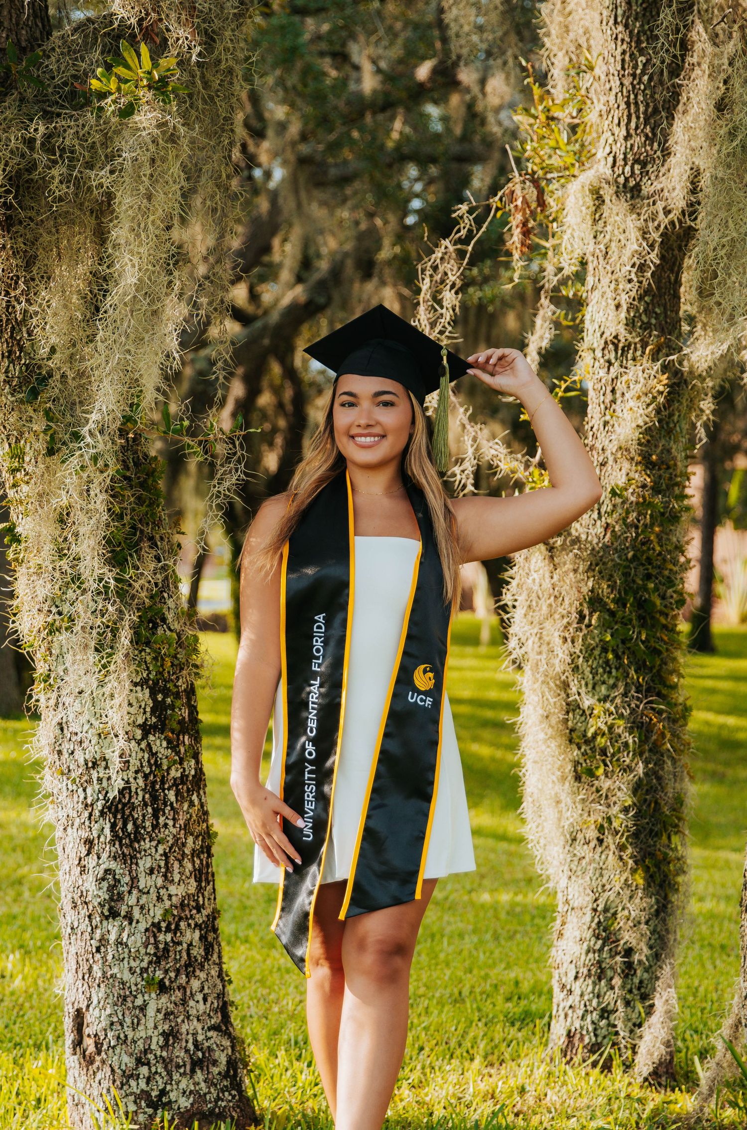 Graduate in black cap and gown poses joyfully among Spanish moss-draped trees during outdoor photo session.