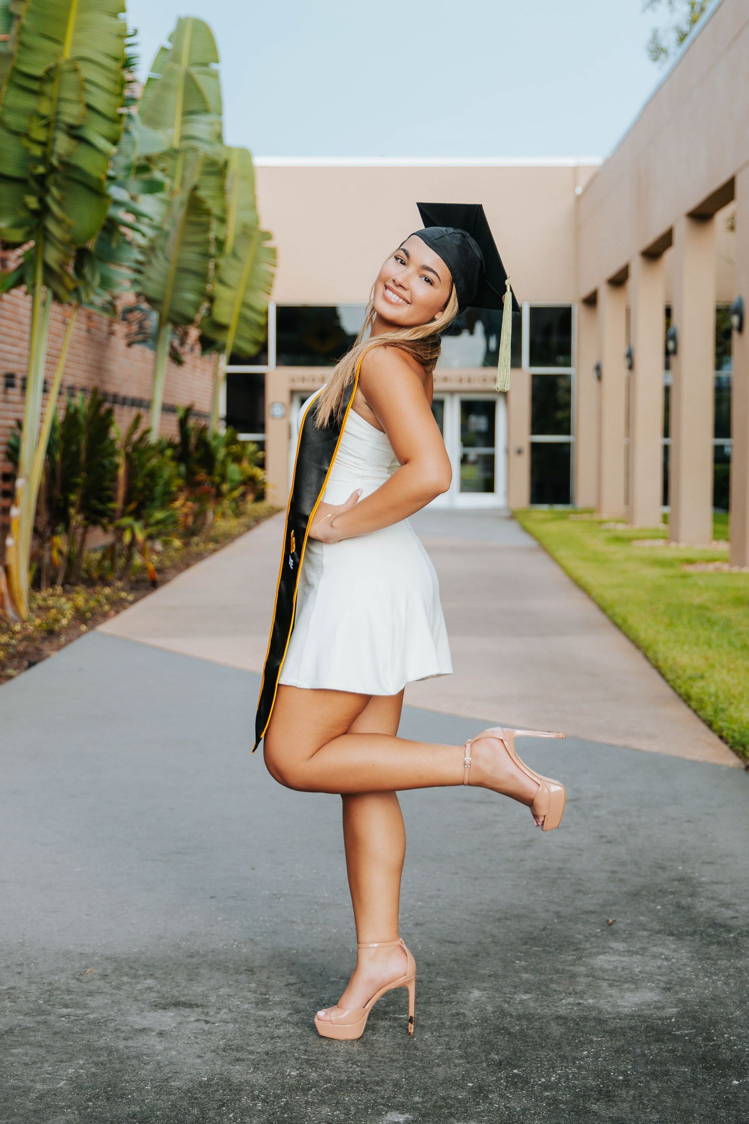 Graduate in white dress and heels poses elegantly on campus walkway surrounded by tropical plants.