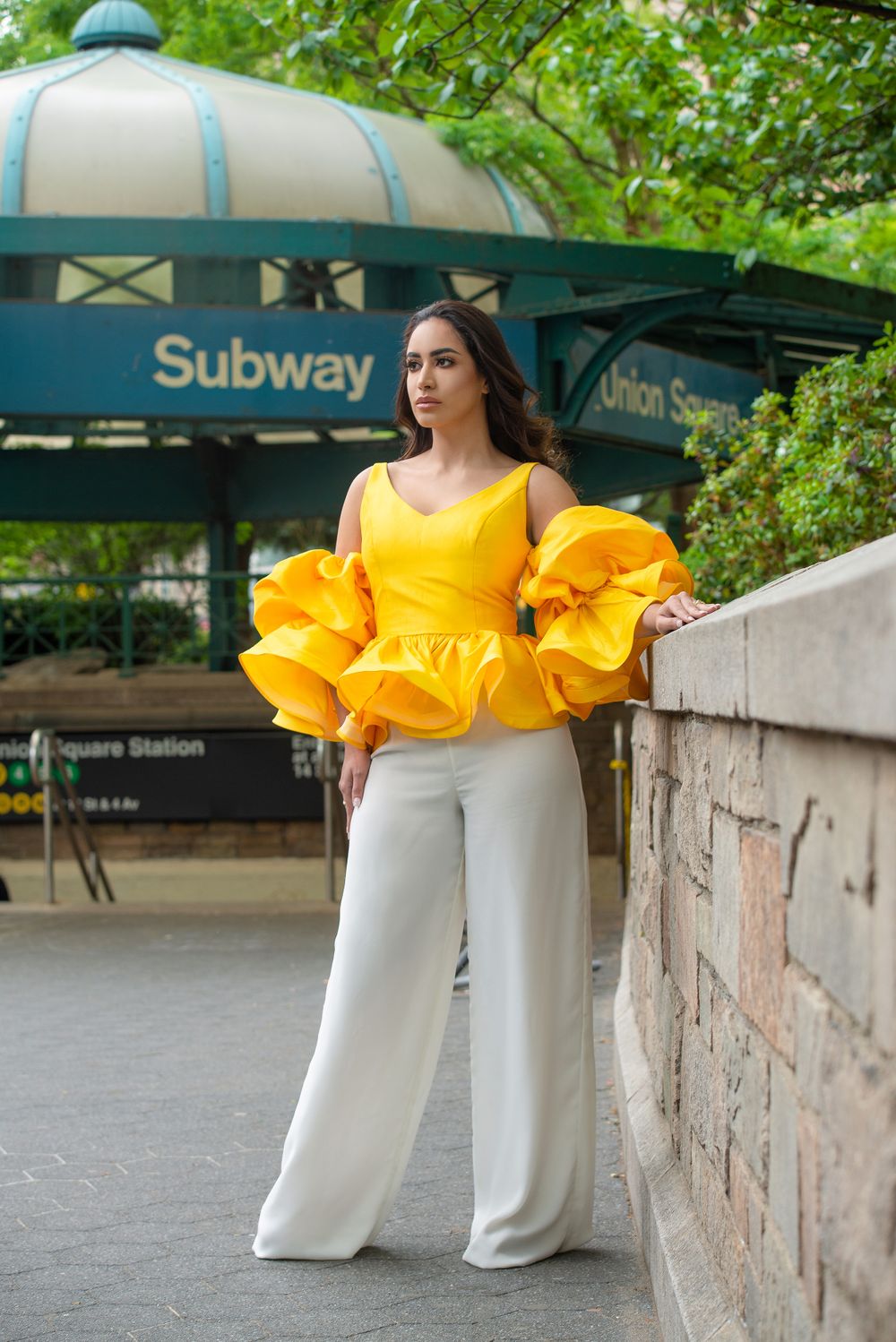 Woman in dramatic yellow ruffled top and white pants poses at NYC subway entrance.