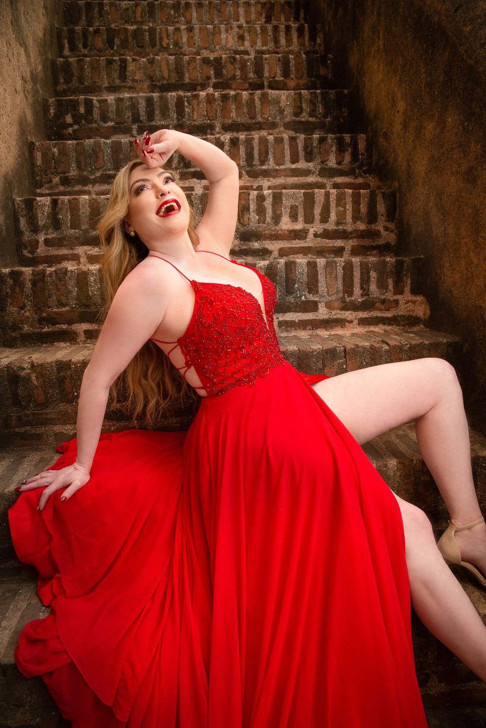 Glamorous red evening gown photo against rustic brick stairs with dramatic pose.