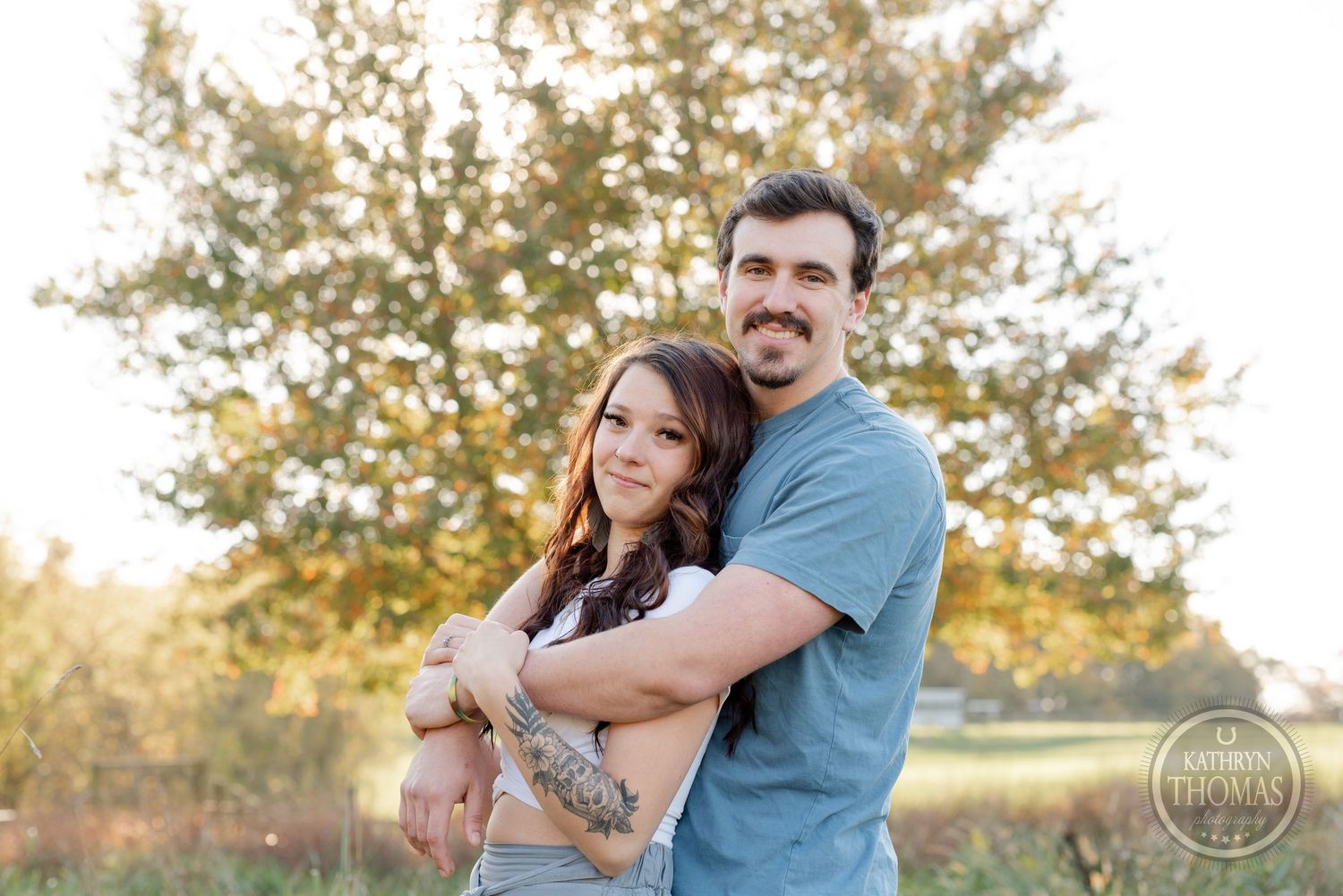 A couple poses together in casual attire against a backdrop of autumn trees.