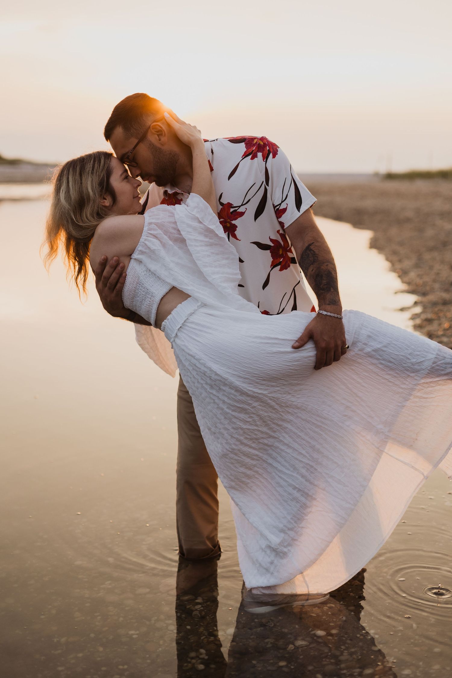 Romantic couple embracing at sunset on a beach with flowing white dress and floral shirt.
