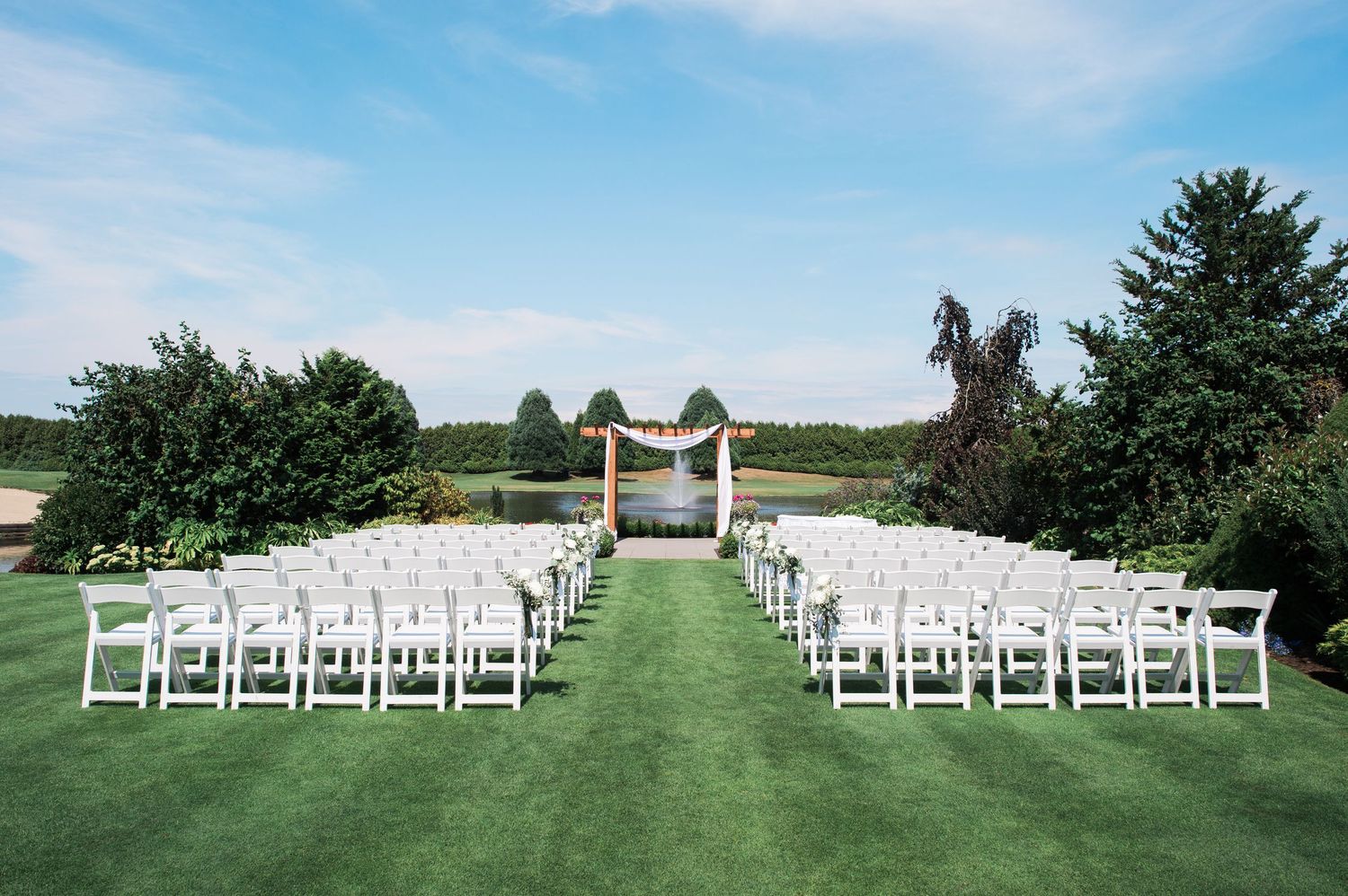 Outdoor wedding ceremony setup with white chairs and wooden arch on manicured lawn.