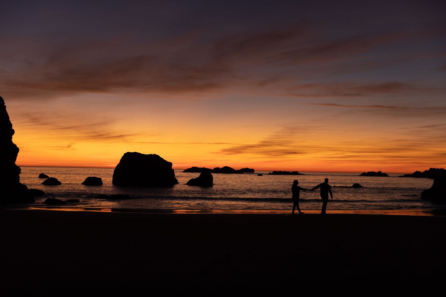 Silhouettes walking on beach at sunset with dramatic orange sky and rock formations.