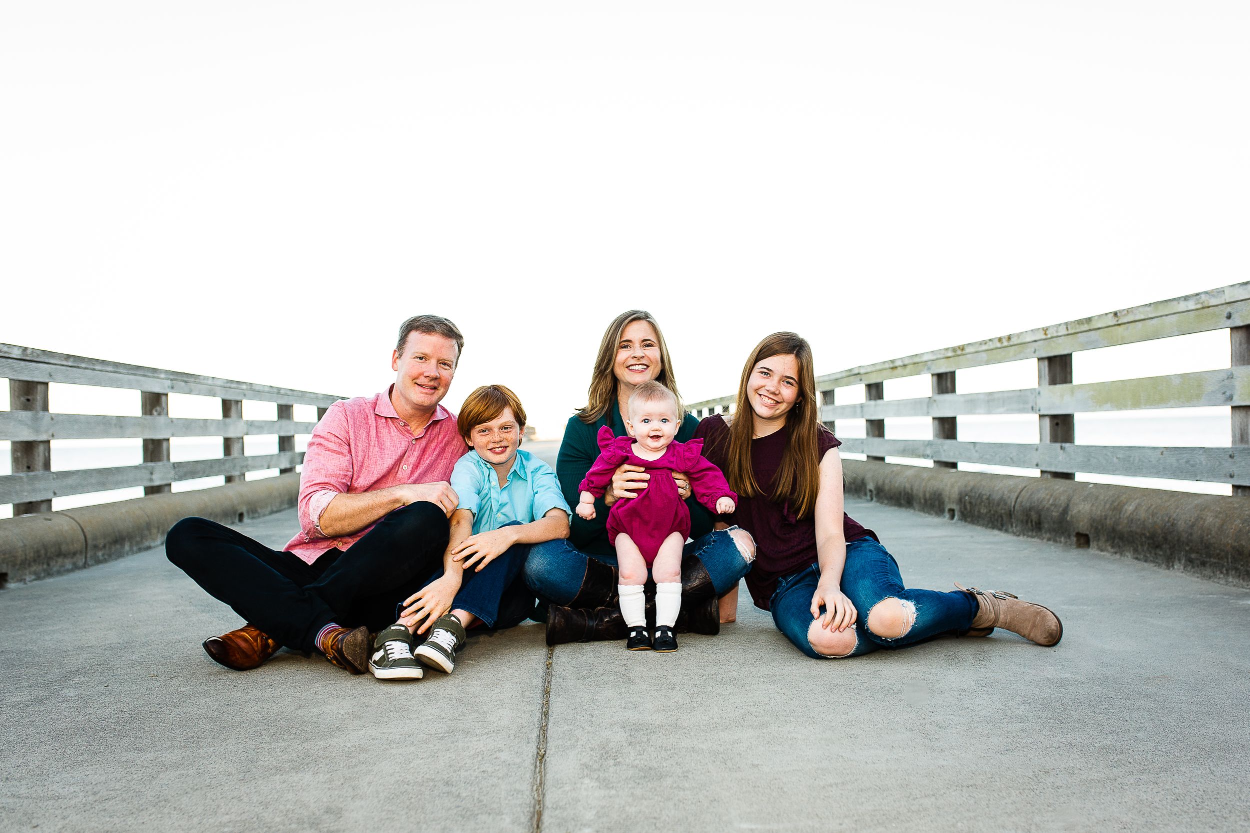 Happy family of five sitting together on a wooden bridge, smiling outdoors in a bright, open setting.