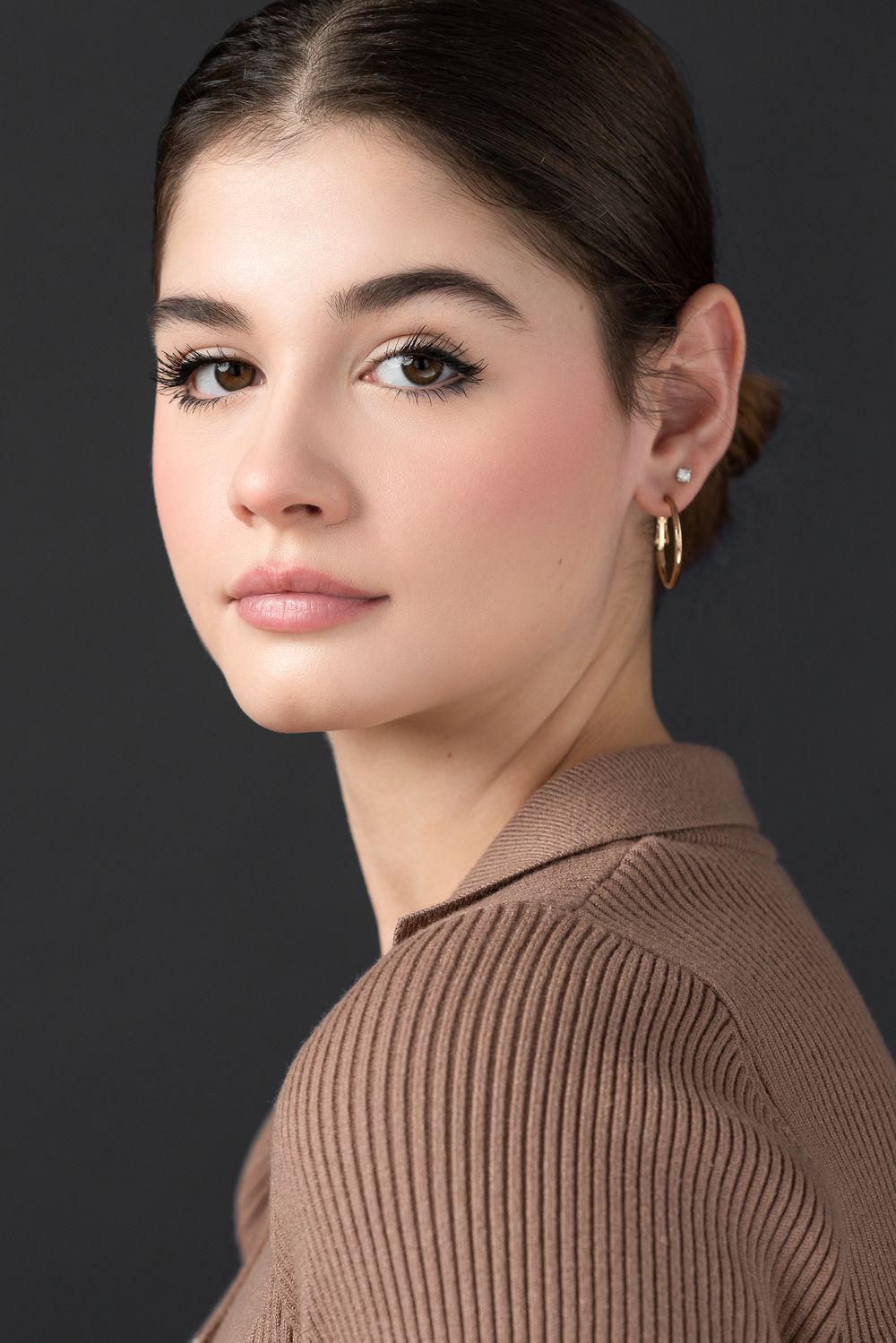 Professional headshot against dark background featuring brown ribbed sweater and gold hoop earrings.