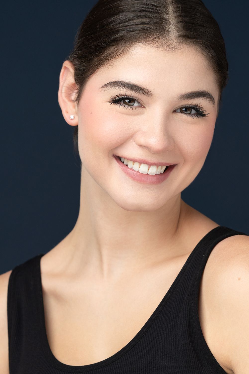Studio portrait in black tank top with natural makeup and bright smile against navy background.