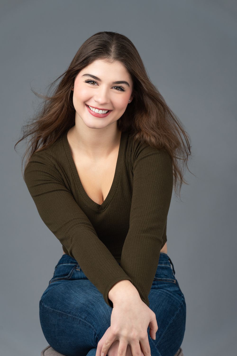 Casual portrait in olive sweater and jeans with flowing brown hair against grey background.