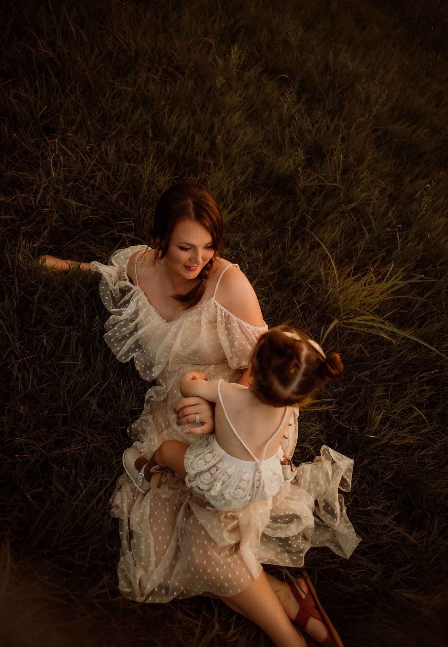 Two figures in white lace dresses share an intimate moment in tall grass at dusk.
