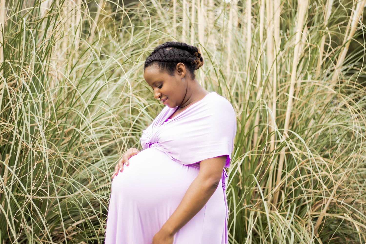 Expecting mother in pink dress standing among tall grass during outdoor maternity shoot.