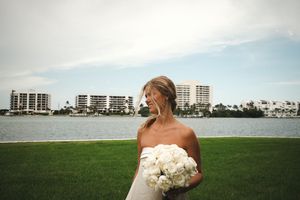 Bride in strapless gown holding white peony bouquet stands on green lawn with waterfront buildings in background.